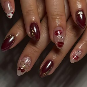 May include: A set of manicured nails featuring a deep burgundy color with gold accents and delicate designs. Some nails showcase clear bases with red lip and bow designs, while others have gold bow and cherry designs. The nails are oval-shaped.