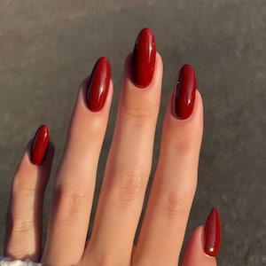 May include: A hand with long, oval-shaped nails painted a deep, glossy red. The nails are evenly shaped and reflect light, creating a polished and elegant appearance. The background is a neutral gray.