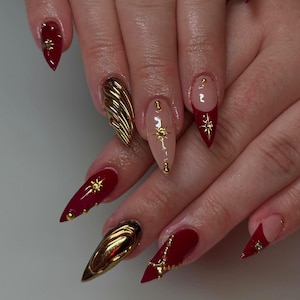 Pode incluir: Close-up de uma mão com unhas stiletto vermelhas e nude decoradas com detalhes dourados, incluindo estrelas, correntes e um design de concha dourada em 3D.
