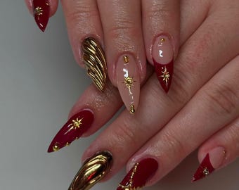Puede incluir: Primer plano de una mano con uñas de stiletto rojas y nude decoradas con detalles dorados, incluyendo estrellas, cadenas y un diseño de concha dorada en 3D.
