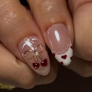 May include: Close-up of manicured nails with a light pink base. One nail features a gold bow with red cherries, and a white design. Another nail has a white tip with red hearts and a scalloped edge. Nail art.