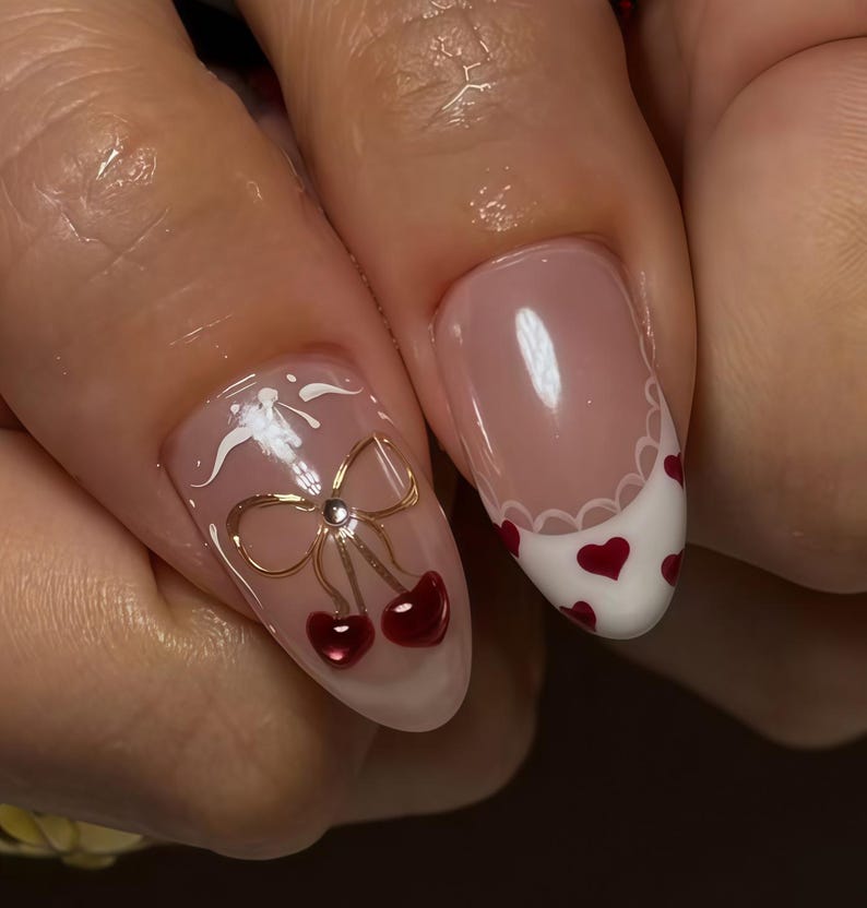 May include: Close-up of manicured nails with a light pink base. One nail features a gold bow with red cherries, and a white design. Another nail has a white tip with red hearts and a scalloped edge. Nail art.
