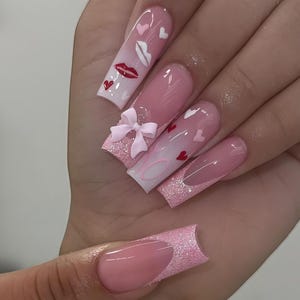 May include: Long, square-shaped artificial nails with a pink and white gradient design. The nails feature red lips, hearts, and a pink bow. Some nails have a glittery pink base and a French tip design.
