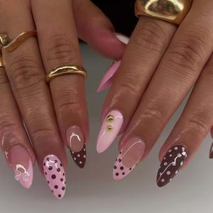May include: A close-up of a hand with long, almond-shaped nails. The nails feature a variety of designs, including pink with polka dots, brown with white polka dots, and a French tip design. Gold rings adorn the fingers.