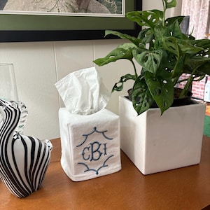 May include: A white square tissue box cover with blue embroidered initials and a decorative border. A green potted plant in a white square pot sits next to the tissue box. A zebra figurine is on the left.