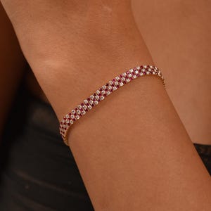 Puede incluir: Una pulsera dorada con un diseño de tres filas. La pulsera está adornada con filas alternas de pequeñas piedras preciosas redondas rosas y transparentes. La joya se lleva en una muñeca.