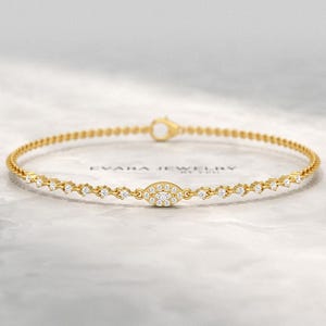 May include: A delicate gold chain bracelet featuring a series of small, round, clear stones set along the band. The centre of the bracelet showcases an oval-shaped charm encrusted with multiple tiny clear stones. The text EVARA JEWELRY is visible.