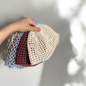 May include: A hand holds a stack of four crochet bucket hats in various colors. The hats are light beige, burgundy, and light blue. The hats have a large open weave design.