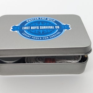 May include: A silver metal tin with a blue and white logo that says "Master the Wild Lost Boys Survival Co. Covert Tools for Survival". The tin is open and contains a roll of white cord and a small silver container.