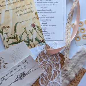May include: A collection of craft supplies including printed cards with text, floral illustrations, and a sketch. There is a cream-colored lace ribbon, a pink ribbon, and a piece of tan fabric with a floral pattern. The text includes Bible verses and prompts for observation.