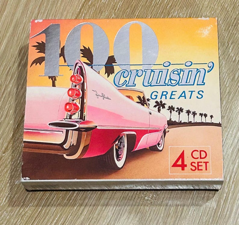 Może przedstawiać: Zestaw płyt CD zatytułowany "100 Cruisin' Greats" z ilustracją r&oacute;żowego zabytkowego samochodu. Okładka albumu ma retro design z palmami i tłem zachodu słońca. Zestaw zawiera 4 płyty CD.