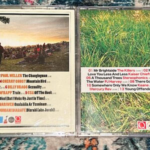 Może przedstawiać: Otwarty futerał na płyty CD, prezentujący okładkę albumu i listy utwor&oacute;w. Lewa strona przedstawia scenę koncertową o zachodzie słońca, a prawa strona pole zielonej trawy. Listy utwor&oacute;w są wydrukowane białym tekstem.
