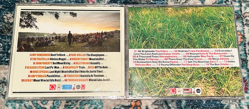 Może przedstawiać: Otwarty futerał na płyty CD, prezentujący okładkę albumu i listy utwor&oacute;w. Lewa strona przedstawia scenę koncertową o zachodzie słońca, a prawa strona pole zielonej trawy. Listy utwor&oacute;w są wydrukowane białym tekstem.