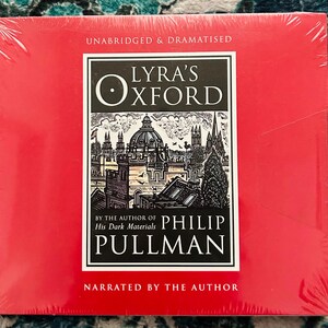 May include: A red, shrink-wrapped audiobook of "Lyra's Oxford" by Philip Pullman. The cover features a black and white illustration of a city with the title in a white and black box. Text on the cover reads "Unabridged & Dramatised" and "Narrated by the Author."
