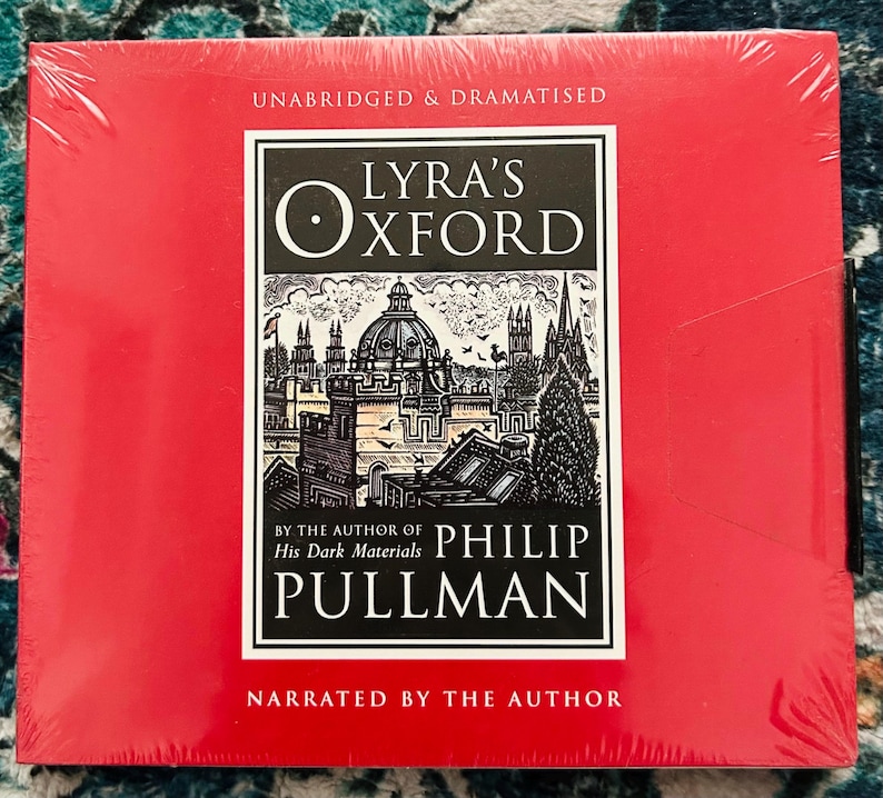 May include: A red, shrink-wrapped audiobook of "Lyra's Oxford" by Philip Pullman. The cover features a black and white illustration of a city with the title in a white and black box. Text on the cover reads "Unabridged & Dramatised" and "Narrated by the Author."