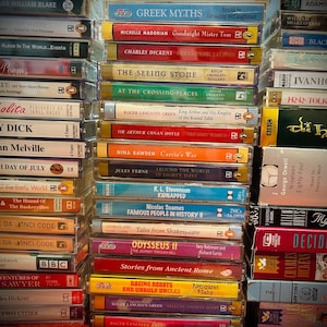 Könnte beinhalten: Mehrere Stapel alter Audiokassetten. Titel wie "Moby Dick", "Greek Myths" und "Nineteen Eighty-Four" sind zu sehen. Die Kassetten zeigen farbenfrohe Etiketten und Texte, die eine Vielzahl literarischer Werke darstellen.
