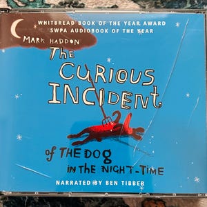 May include: Audiobook CD of "The Curious Incident of the Dog in the Night-Time" by Mark Haddon. The cover is blue with white text and a red dog illustration. The text includes "WHITBREAD BOOK OF THE YEAR AWARD" and "SWPA AUDIOBOOK OF THE YEAR".