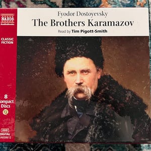 May include: A boxed set of "The Brothers Karamazov" audiobook by Fyodor Dostoyevsky, read by Tim Pigott-Smith. The cover features a portrait of a man with a large mustache. The box is red and white with the Naxos Audiobooks logo and the text "Classic Fiction".
