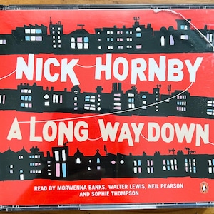 May include: A CD case with a red cover featuring the title "A Long Way Down" by Nick Hornby. The cover art depicts a black cityscape silhouette against a red background. The text is white. The CD case is in a clear plastic case.