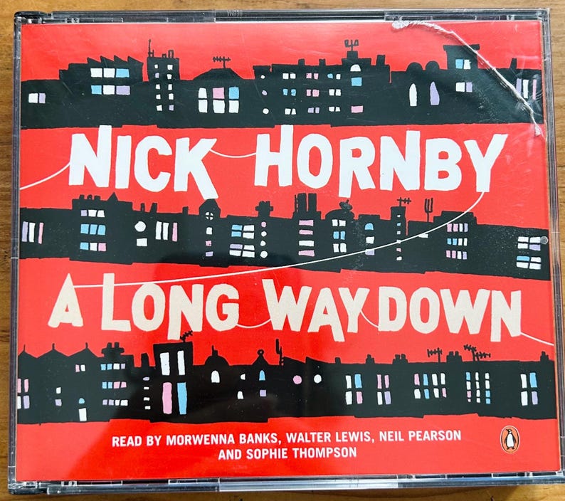 May include: A CD case with a red cover featuring the title "A Long Way Down" by Nick Hornby. The cover art depicts a black cityscape silhouette against a red background. The text is white. The CD case is in a clear plastic case.