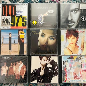 Może przedstawiać: Wyświetlono dziewięć album&oacute;w muzycznych na CD, z r&oacute;żnymi artystami. Okładki album&oacute;w to m.in. "Old 97's", "Lonely at the Top", "Older", "Sinead O'Connor", "Patty Smyth", "Maroon 5", "Prince" i "Jess Nicholls - Sings". Płyty CD są w przezroczystych plastikowych etui.