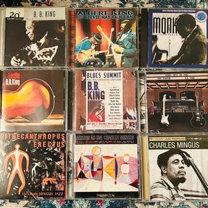Puede incluir: Una colección de nueve CD de música con artistas de blues y jazz. Los CD muestran portadas de álbumes con imágenes de B.B. King, Albert King, Stevie Ray Vaughan, Thelonious Monk y Charles Mingus. Las portadas tienen texto.