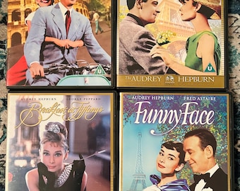 May include: Four DVD movie cases featuring Audrey Hepburn. Titles include "Roman Holiday," "Paris When It Sizzles," "Breakfast at Tiffany's," and "Funny Face." Each case has a gold border and features movie stills of the actors.