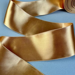 Puede incluir: Un rollo de cinta de raso dorada, parcialmente desenrollada, descansa sobre una superficie azul claro. La cinta tiene una textura suave y brillante con un brillo sutil. Los bordes de la cinta están cuidadosamente acabados con una costura fina.