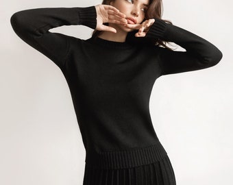 Pode incluir: Conjunto preto de camisola e saia plissada. A camisola de manga comprida tem gola, punhos e bainha canelados. A saia é plissada. A pessoa tem cabelo longo e escuro.