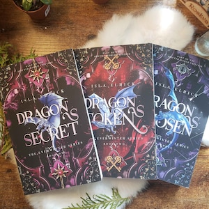 May include: Three books from the Everwinter Series are displayed. The covers feature dragon illustrations and titles: "Dragon Secret," "Dragon Token," and "Dragon Chosen." The books have ornate, dark backgrounds with key motifs and the author's name, Isla Elrick.