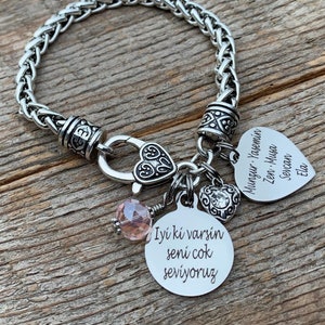 May include: A silver chain bracelet with multiple charms. The charms include a heart with the text "İyi ki varsın seni çok seviyoruz", a heart with the text "Mungur - Yasemin Zen - Musa Sevan Ela", and a heart with a rhinestone.