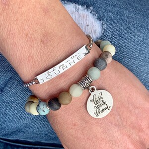 May include: A silver bracelet with a charm that says "live your dream" and a bar charm that says "find joy in the journey". The bracelet is made of beads in shades of brown, gray, and green.