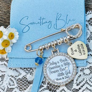 May include: A gold-tone safety pin brooch with clear rhinestones, a blue crystal bead, and a heart-shaped charm engraved with "Evan & Laikelyn 9-26-2026". A round pendant features the text "Today a Bride, Tomorrow a Wife, Forever Our Little Girl. Mom & Dad".