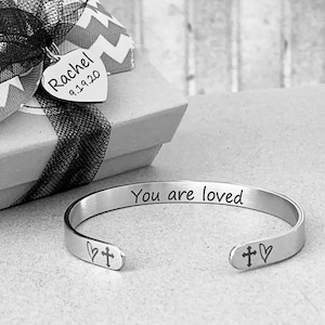 May include: A silver bracelet with the words "You are loved" engraved on it. The bracelet has a heart and cross design on each end.