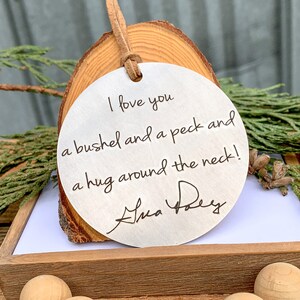 May include: A round, silver ornament with a wood slice backing. The ornament has the text "I love you a bushel and a peck and a hug around the neck!" written in cursive. The ornament is signed "Ana Frey".