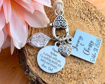May include: A silver charm bracelet with a heart charm, a clear crystal charm, and a silver tag engraved with the text "Mommy & Chrissy October 31, 2020". The bracelet is on a wooden surface with a pink flower.