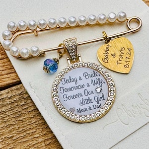 May include: A gold safety pin with a pearl chain, a heart charm with the text "Gabby & Travis 8.17.24", and a round charm with rhinestones and the text "Today a Bride, Tomorrow a Wife, Forever Our Little Girl Mom & Dad".
