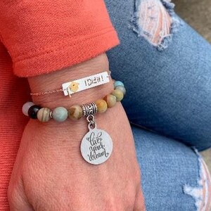 May include: A beaded bracelet with a charm that says "Live your dream" and a silver bar charm that says "I Did It!" The bracelet is made of various colors of stones, including brown, green, and blue.