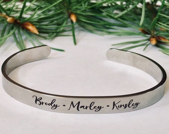 May include: A silver-colored cuff bracelet with the names "Brody", "Marley", and "Kinsley" engraved in a delicate script. The bracelet is set against a backdrop of green pine needles.