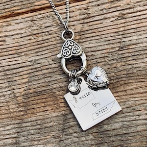 May include: A silver necklace with a heart-shaped charm and a charm with the outline of Washington state. The Washington state charm has the coordinates 99110 and 37135 and the text "'s" inside a heart.