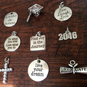 May include: A collection of silver-toned charms with various inspirational quotes and symbols. Charms include a graduation cap, a cross, and the year 2016. The charms are displayed on a dark wooden surface.