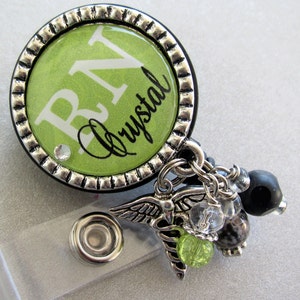 May include: A green and silver badge reel with a green background and white text that reads "RN Crystal". The reel has a silver chain with a silver charm of a caduceus and other charms.