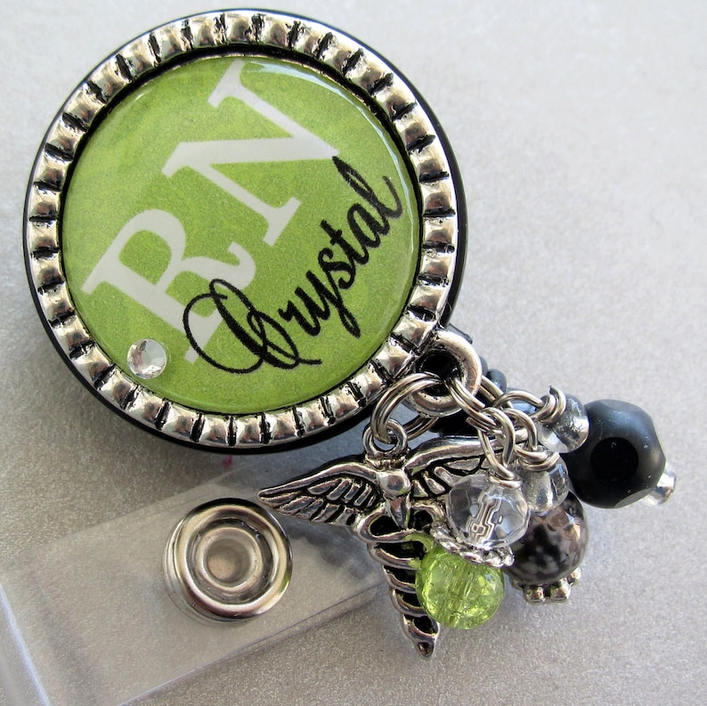 May include: A green and silver badge reel with a green background and white text that reads "RN Crystal". The reel has a silver chain with a silver charm of a caduceus and other charms.