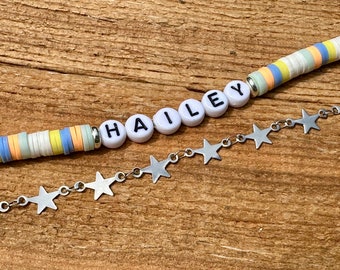 May include: A white beaded bracelet with the name "HAILEY" spelled out in black letters. The bracelet is paired with a silver chain with star-shaped charms.