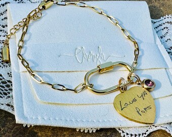 May include: Gold-toned bracelet with a carabiner clasp and a heart-shaped charm. The charm has the handwritten text "Love Ya Pops" and a small pink gemstone. The bracelet is on a white pouch.