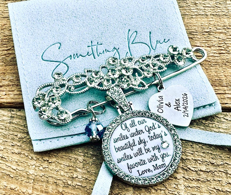 May include: A silver-tone safety pin brooch adorned with clear crystals, featuring a round pendant with the text "Of all our smiles under God's beautiful sky, today's smiles will be my favorite with you. Love, Mom" and a heart charm engraved "Olivia & Alex 2/14/2026".
