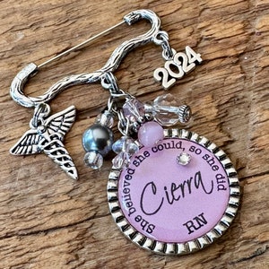 May include: Silver-toned brooch with a safety pin closure. The brooch features a charm with the name "Cierra" and the letters "RN" on a lavender background, surrounded by the phrase "she believed she could, so she did." Additional charms include a caduceus, beads, and the year "2024."