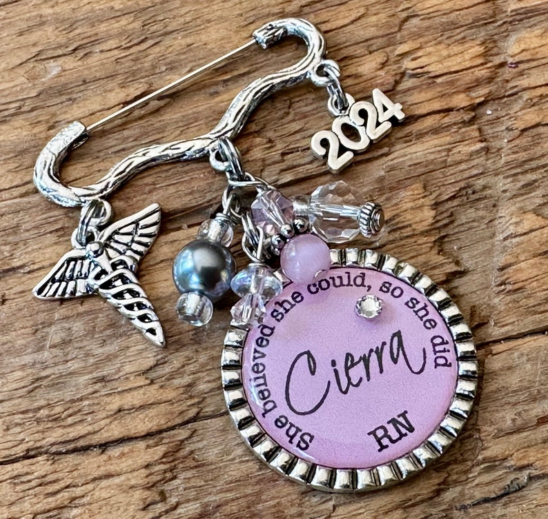 May include: Silver-toned brooch with a safety pin closure. The brooch features a charm with the name "Cierra" and the letters "RN" on a lavender background, surrounded by the phrase "she believed she could, so she did." Additional charms include a caduceus, beads, and the year "2024."