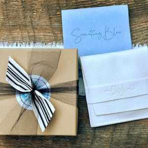 May include: A square brown cardboard gift box tied with a black and white striped ribbon, a light blue square pouch with the text Something Blue, and a white square pouch with a debossed logo, all arranged together on a wooden surface.