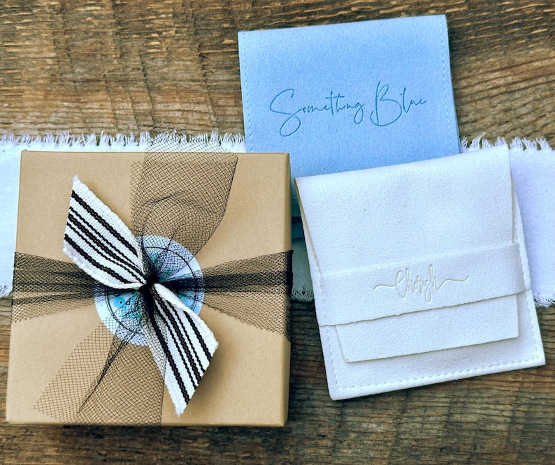 May include: A square brown cardboard gift box tied with a black and white striped ribbon, a light blue square pouch with the text Something Blue, and a white square pouch with a debossed logo, all arranged together on a wooden surface.